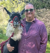 photo of PJ wearing sunglasses and side hugging a black and white llama wearing a red halter